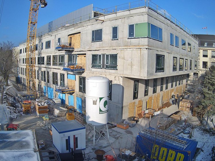 Mehrstöckiges Gebäude im Rohbau mit Betonfassade und bereits eingesetzten Fenstern, umgeben von einer Baustelle mit Kran, Baumaterialien und Containern. Vor dem Gebäude stehen Baugeräte und ein großer zylindrischer Silobehälter, während im Hintergrund Bäume und weitere Gebäude zu sehen sind.