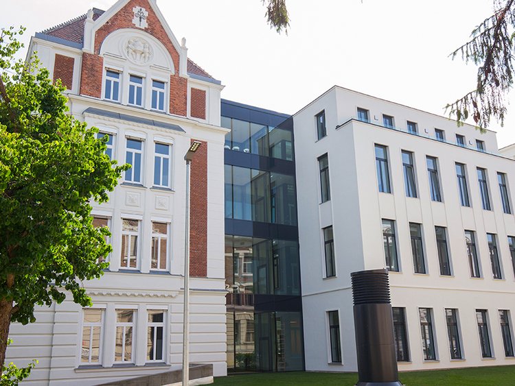 Ein historisches mehrstöckiges Gebäude mit weißer Fassade und roten Backsteinelementen ist durch einen verglasten Übergang mit einem modernen, hellen Anbau, verbunden. Im Vordergrund befinden sich Rasenflächen und Bäume, vor dem Gebäude ragt eine Entlüftungsanlage aus dem Boden.