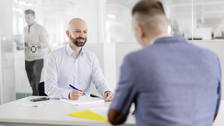 Zwei Personen sitzen sich an einem Schreibtisch in einem Büro gegenüber und führen ein Gespräch. Eine Person hält einen Stift über Unterlagen, während im Hintergrund durch eine Glaswand eine weitere Person zu sehen ist.