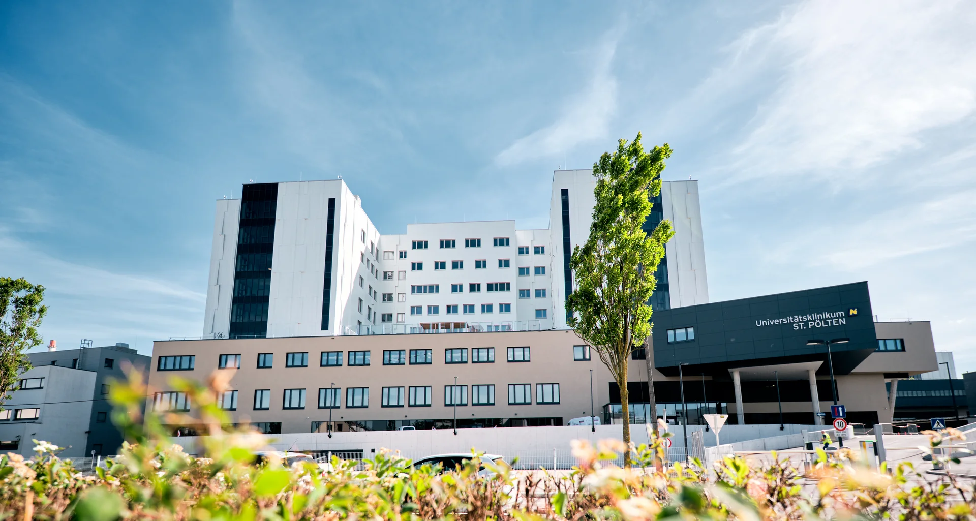 Modernes mehrstöckiges Krankenhausgebäude mit hellem Fassadenmix aus Weiß und Beige sowie einem dunklen Eingangsbereich mit Beschriftung. Im Vordergrund befinden sich unscharfe Pflanzen, dahinter führt eine Zufahrt zum Haupteingang, während der Himmel blau mit leichten Wolken ist.