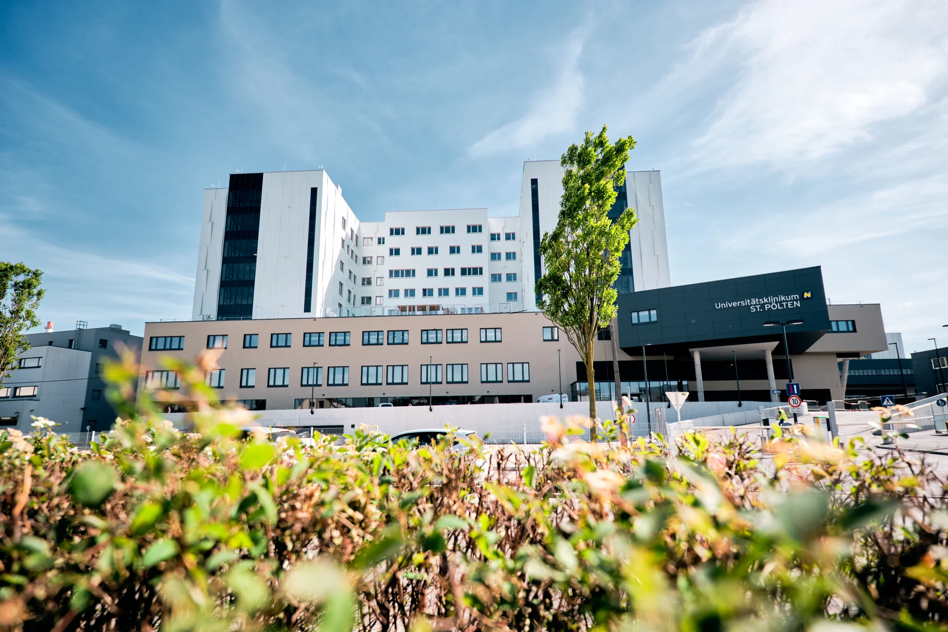 Modernes mehrstöckiges Krankenhausgebäude mit hellem Fassadenmix aus Weiß und Beige sowie einem dunklen Eingangsbereich mit Beschriftung. Im Vordergrund befinden sich unscharfe Pflanzen, dahinter führt eine Zufahrt zum Haupteingang, während der Himmel blau mit leichten Wolken ist.