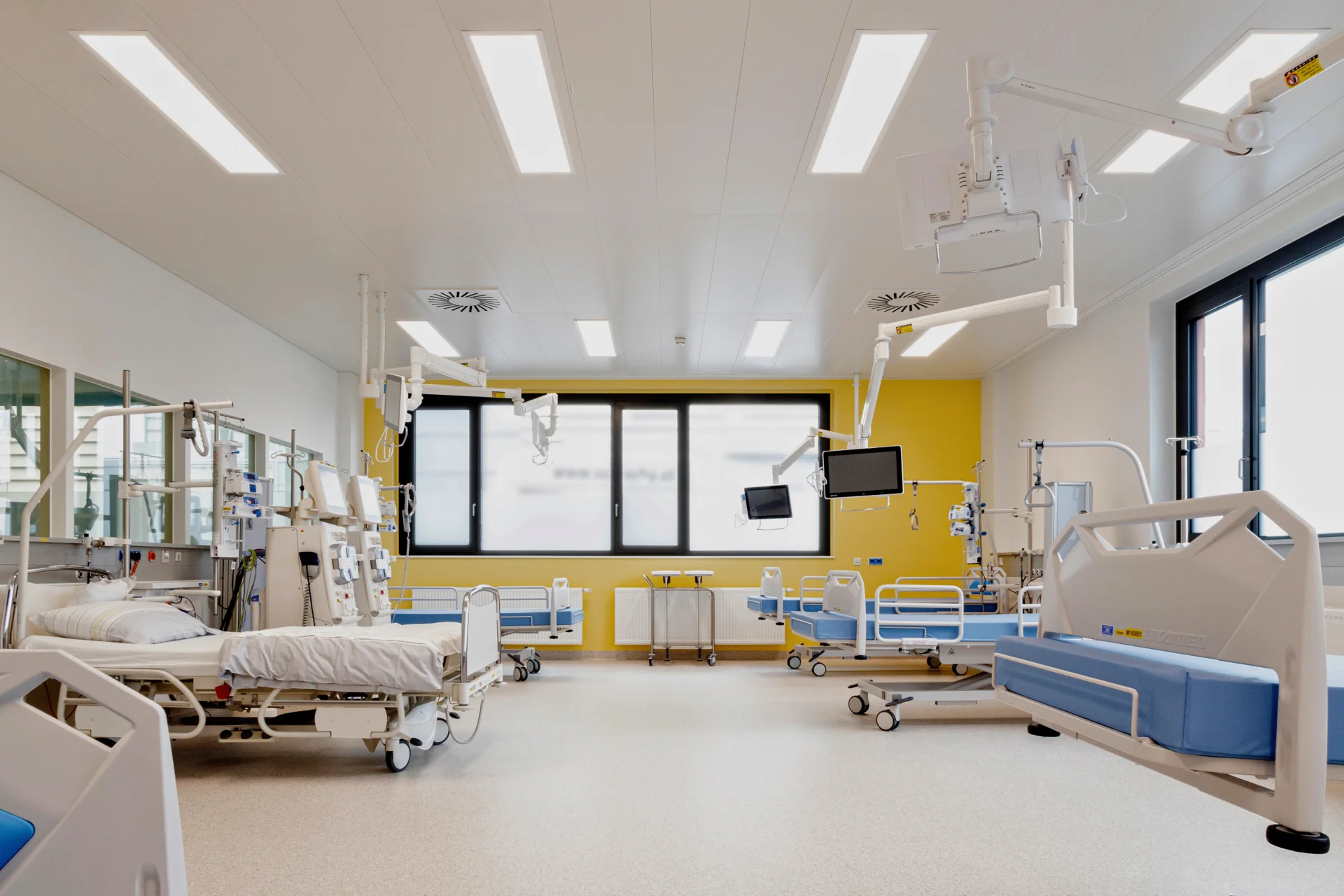 Heller, moderner Krankenhausraum mit mehreren leeren Pflegebetten, medizinischen Geräten an den Betten, Deckenlampen und Monitorarmen. Große Fenster lassen Tageslicht herein; die Wände sind gelb gestrichen, der Boden hell. Keine Personen im Raum.