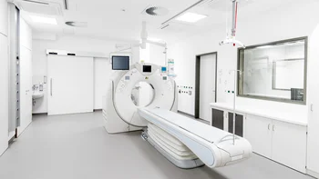 Moderner medizinischer Untersuchungsraum mit einem groĂen CT-Scanner und einer ausfahrbaren Liege in der Mitte. Der Raum ist hell und minimalistisch ausgestattet, mit Monitoren, WandanschlĂŒssen, SchrĂ€nken und einem Sichtfenster zu einem angrenzenden Kontrollraum