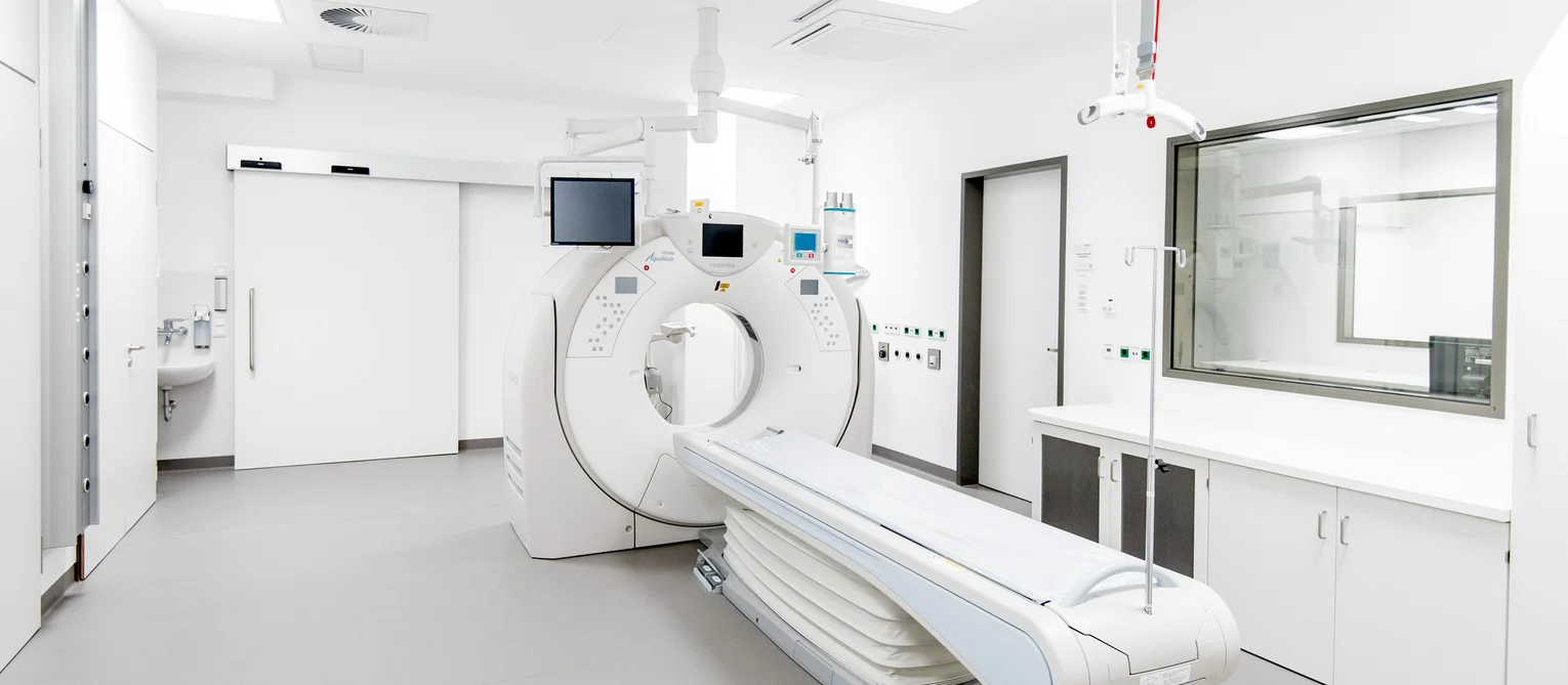 Moderner medizinischer Untersuchungsraum mit einem großen CT-Scanner und einer ausfahrbaren Liege in der Mitte. Der Raum ist hell und minimalistisch ausgestattet, mit Monitoren, Wandanschlüssen, Schränken und einem Sichtfenster zu einem angrenzenden Kontrollraum