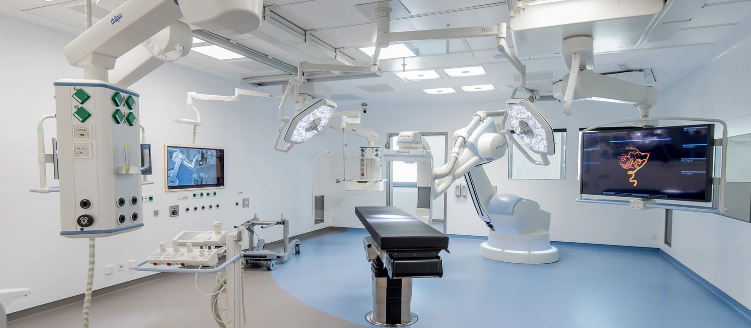 Moderner Operationssaal mit einem zentralen Operationstisch, mehreren Deckenleuchten und medizinischen GerĂ€ten an beweglichen Armen. Rechts steht ein groĂer robotischer OP-Arm neben einem Monitor mit medizinischer Bilddarstellung, wĂ€hrend links zusĂ€tzliche GerĂ€tewagen und Bedienelemente an der Wand angebracht sind.