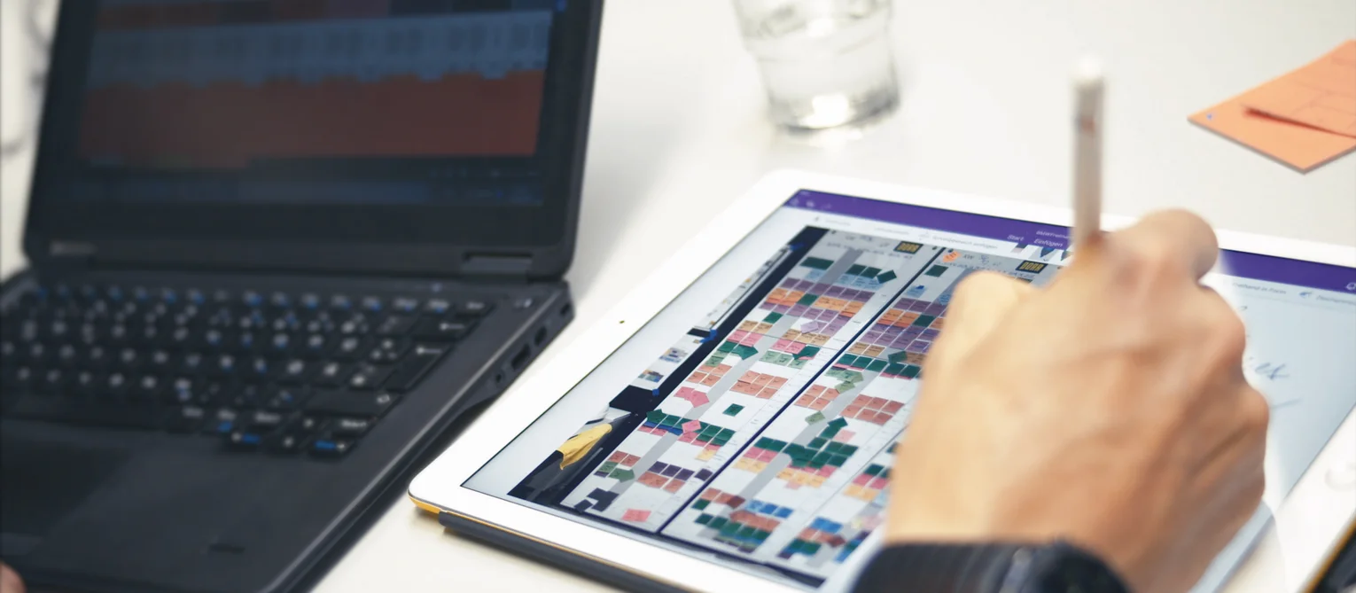 Eine Person hält einen Stift über ein Tablet, auf dem ein farbcodierter digitaler Plan mit vielen kleinen Feldern dargestellt ist. Daneben stehen ein aufgeklappter Laptop und ein Glas Wasser auf einem Tisch.